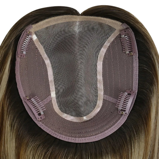 Close-up of the underside of a hairpiece extension, showing a semi-sheer mesh crown, brown adhesive base, and multiple metal hair clips holding the piece in place. The attached synthetic hair is light brown to blonde with subtle warm highlights.