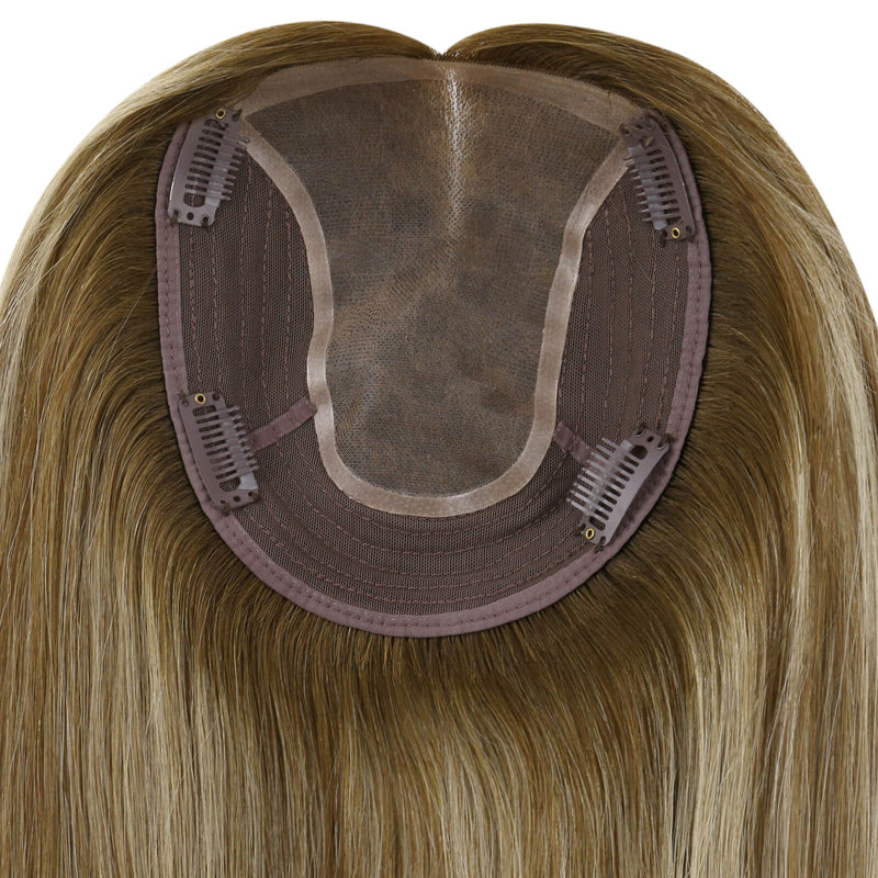 Load image into Gallery viewer, Close-up of the underside of a hairpiece extension, showing a semi-sheer mesh crown, brown adhesive base, and multiple metal hair clips holding the piece in place. The attached human hair is light brown to blonde with subtle warm highlights.
