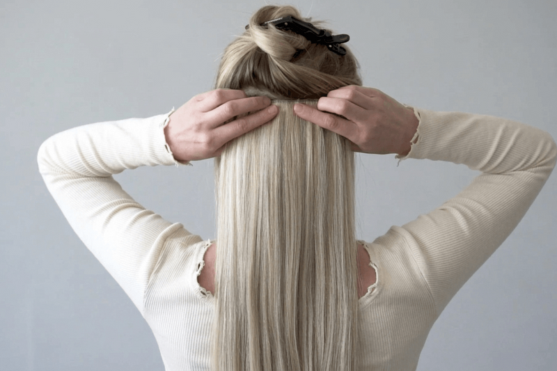 Person with a long blonde wig wearing a beige sweater against a gray background