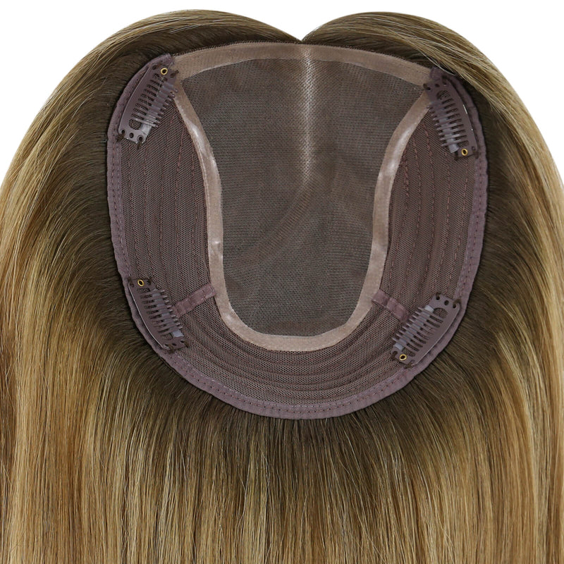 Load image into Gallery viewer, Close-up of the underside of a hairpiece extension, showing a semi-sheer mesh crown, brown adhesive base, and multiple metal hair clips holding the piece in place. The attached synthetic hair is light brown to blonde with subtle warm highlights.
