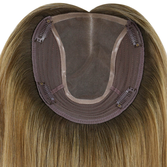 Close-up of the underside of a hairpiece extension, showing a semi-sheer mesh crown, brown adhesive base, and multiple metal hair clips holding the piece in place. The attached synthetic hair is light brown to blonde with subtle warm highlights.