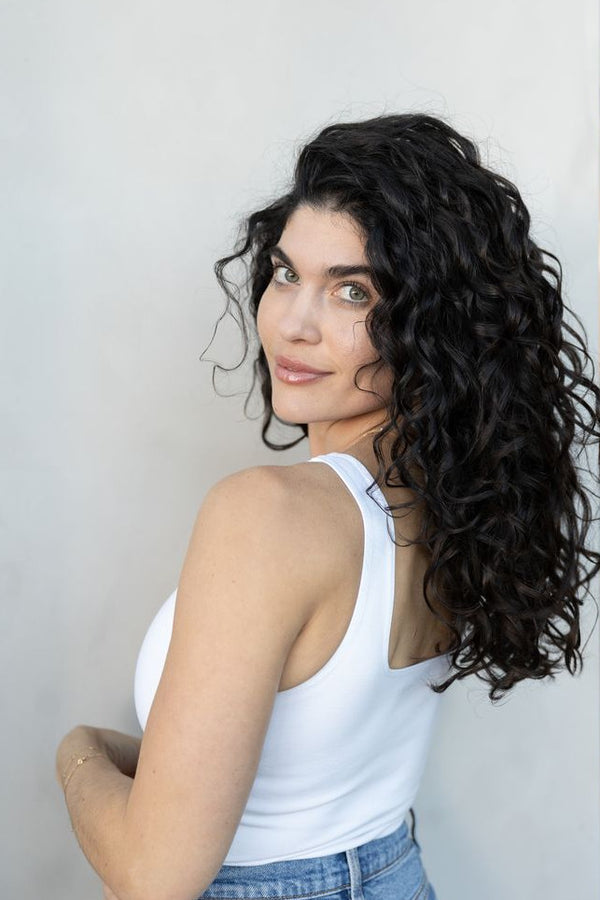 Woman with curly hair wearing a white tank top and blue jeans against a light gray wall.