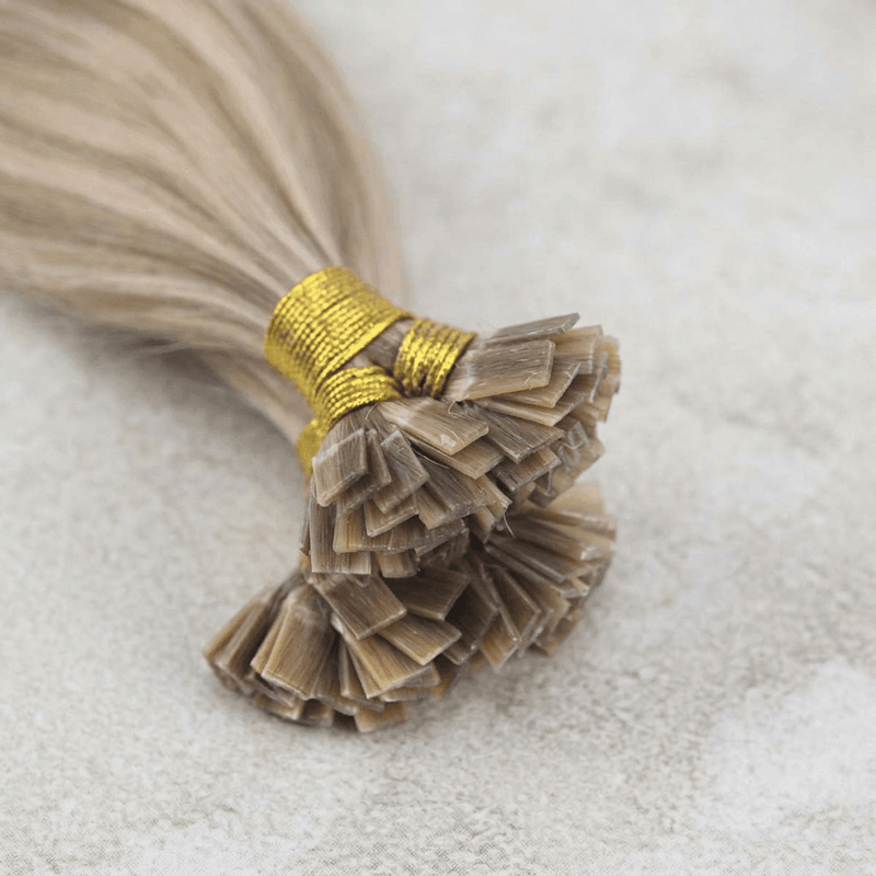 A close-up of a bundle of light blonde or beige hair extensions tied with a gold thread wrap, resting on a soft, light gray textured surface.