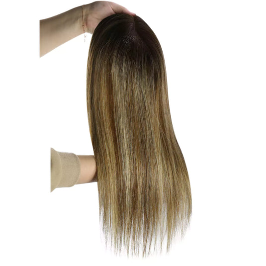 A long hairtopper with a warm golden-brown and light blonde ombré color, held up by a hand against a plain white background.