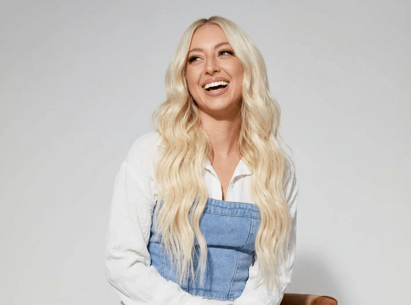 Woman with long blonde hair wearing a denim overalls and white shirt against a plain background