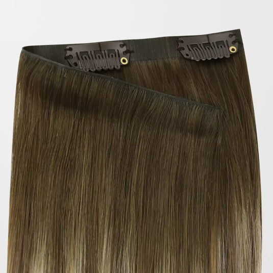 A pack of clip-in hair extensions with straight, medium brown hair featuring subtle warm blonde highlights, attached to black elastic bands with gold metal grommets.