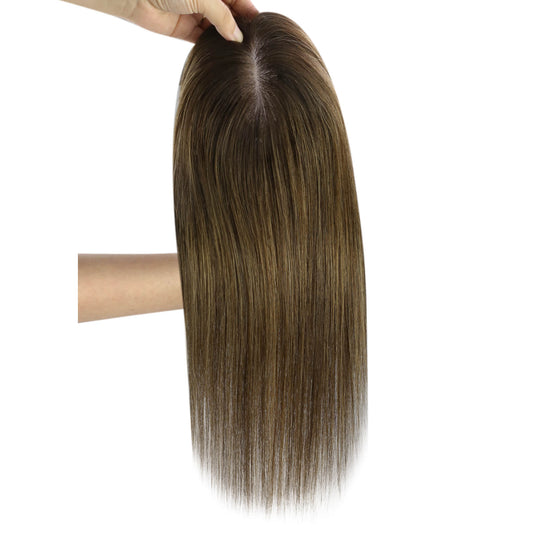 A long hair topper with a dark brown and blonde ombré color, held up by a hand against a plain white background.
