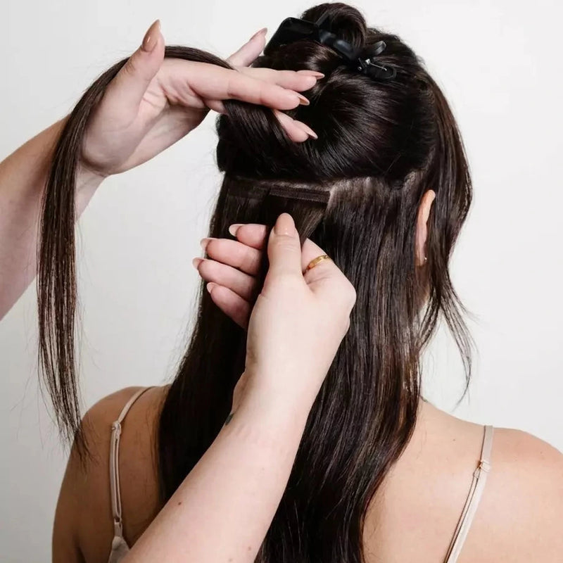 Hairstylist installing tape hair extensions on long dark hair against a plain background