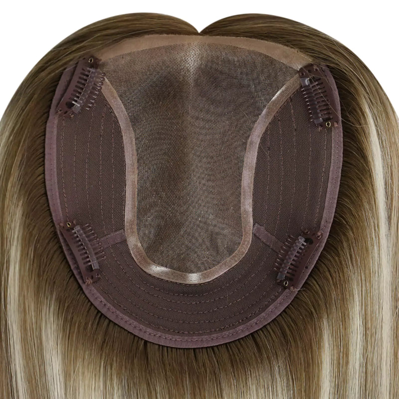 Load image into Gallery viewer, Close-up of the underside of a hairpiece extension, showing a semi-sheer mesh crown, brown adhesive base, and multiple metal hair clips holding the piece in place. The attached human hair is light brown to blonde with subtle warm 
